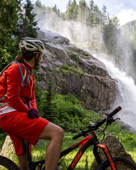 4 Sterne Hotel in Salzburgerland: Smaragdhotel Tauernblick Fahrradfahrerin sitzt und blickt auf einen Wasserfall in einem Wald