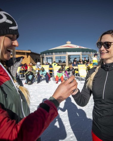 4 Sterne Hotel in Salzburgerland: Smaragdhotel Tauernblick Zwei Frauen stoßen in Skikleidung im Schnee mit Gläsern an, Hintergrund Skihütte und Menschen