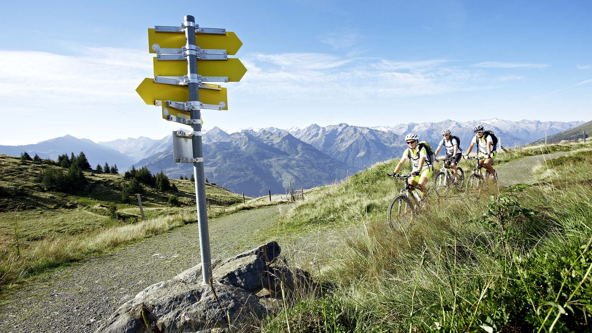 Hohe Tauern National Park: holiday for bikers Three cyclists on mountain trail with hiking signpost and Alps in background