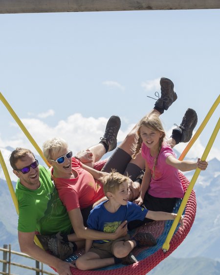 Hotel in Bramberg: Smaragdhotel Tauernblick Familie mit zwei Kindern sitzt lachend auf großer Nestschaukel in den Bergen