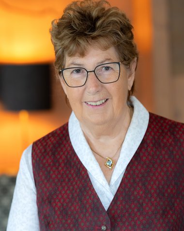 Wildkogel: Hotel mit Blick für das Besondere Ältere Frau mit Brille und roter Weste lächelnd vor warmem Hintergrund