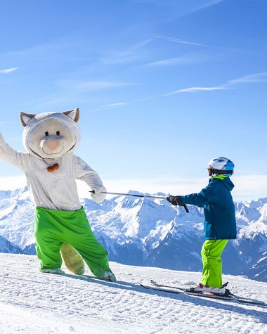 4 Sterne Hotel in Salzburgerland: Smaragdhotel Tauernblick Kind mit blauem Helm hält Skistock einer großen Katze auf Skiern in den Alpen