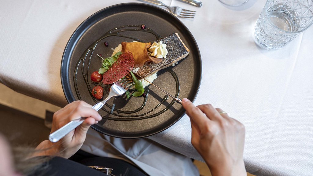 Wellnesshotel Salzburgerland: Smaragdhotel Tauernblick Person schneidet Fischgericht mit Beilagen auf schwarzem Teller an Tisch mit Glas und Besteck