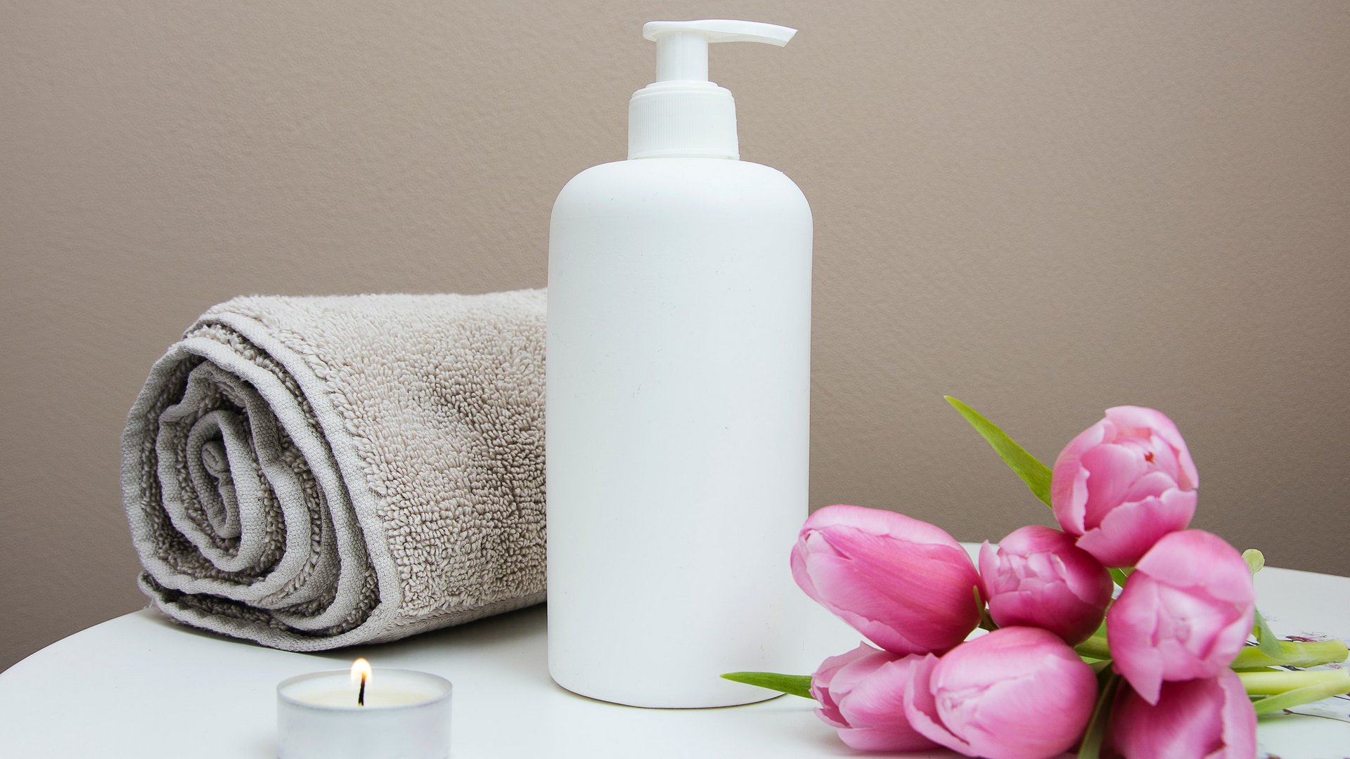 Wasserspaß im Hotel in Österreich mit Pool Weiße Lotionflasche, graues Handtuch, brennende Kerze und rosa Tulpen auf weißem Tisch