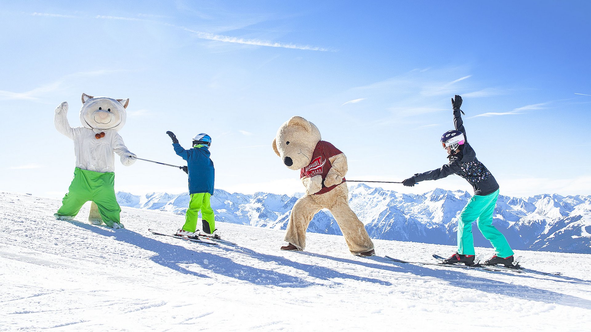 Family hotel in Bramberg: Smaragdhotel Tauernblick Two children on skis pulling two large plush mascots in snow with mountain background