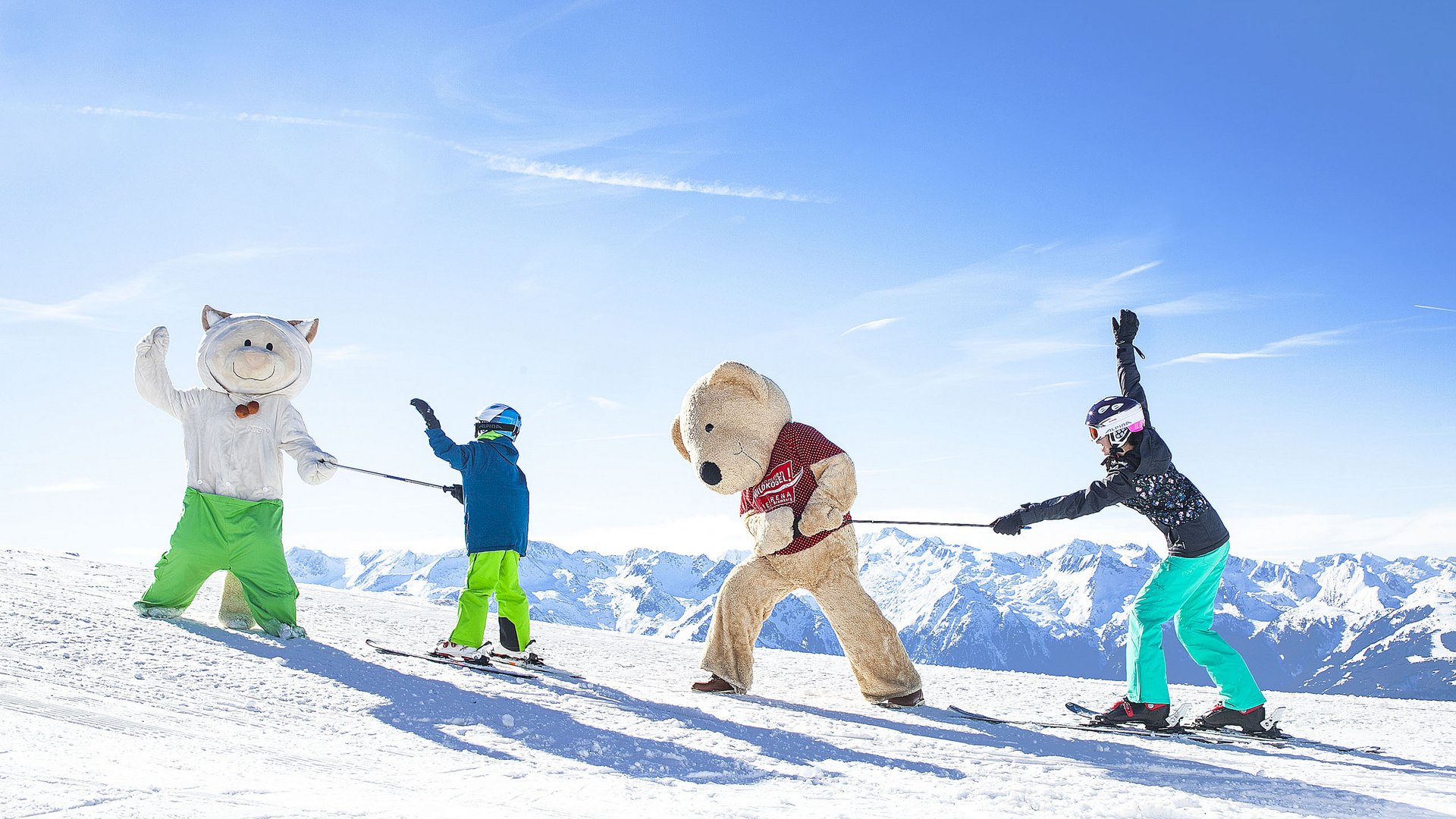 Skiurlaub mit Kindern in Österreich Zwei Kinder auf Skiern ziehen zwei große Plüschfiguren im Schnee vor Bergkulisse