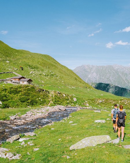 Einmaliges Wanderhotel im Salzburgerland Zwei Wanderer auf grünem Bergweg an Bach mit Almhütte und Alpen im Hintergrund