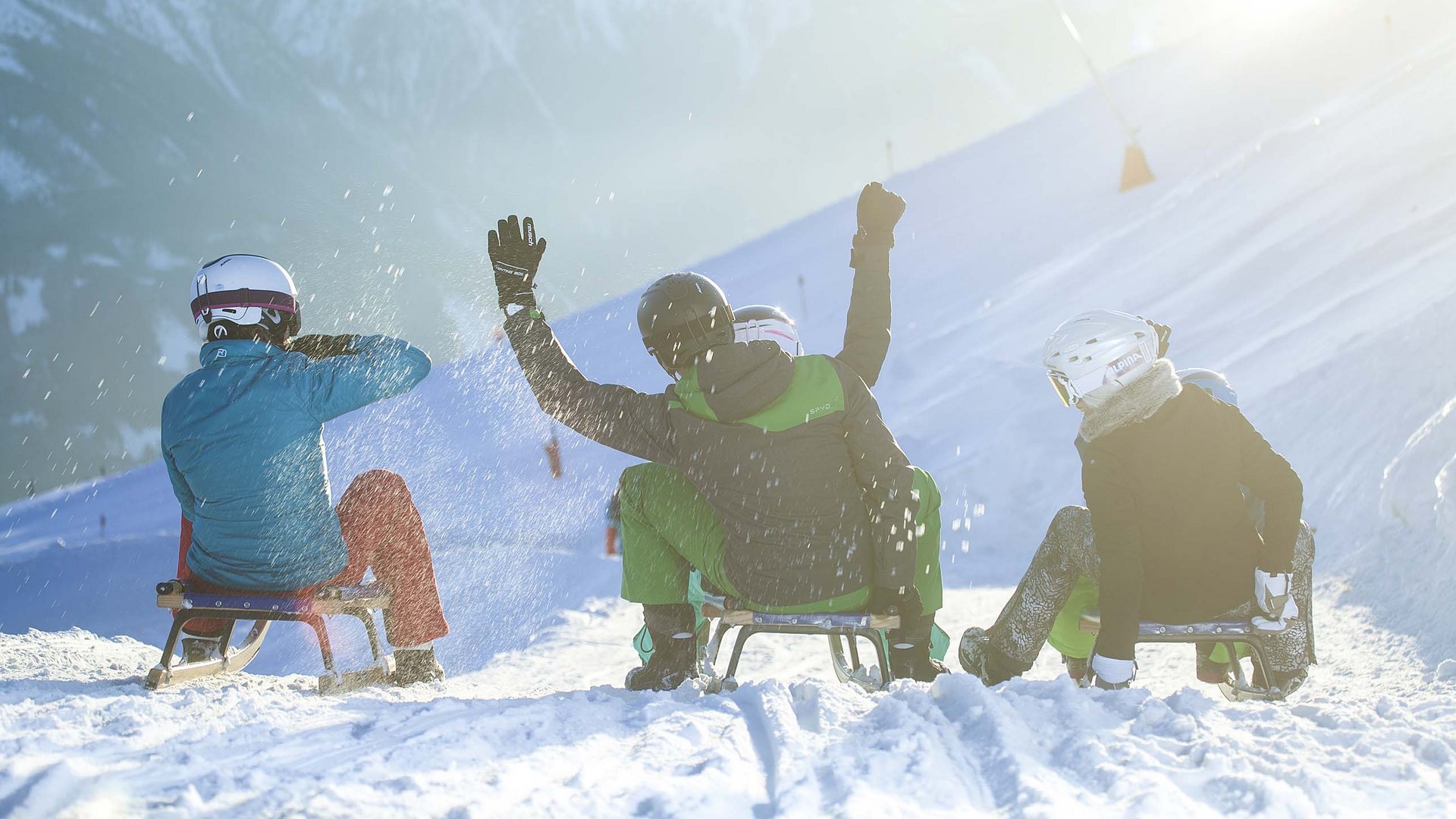 Salzburgerland: Winter im Smaragdhotel Tauernblick Vier Personen rodeln mit Helmen auf verschneiter Piste im Sonnenschein