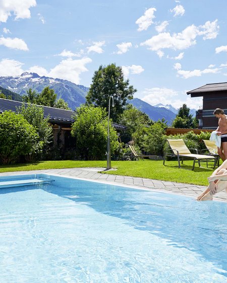 Wellnesshotel Salzburgerland: Smaragdhotel Tauernblick Frau sitzt am Rand eines Pools mit Bergblick und Sonnenschein