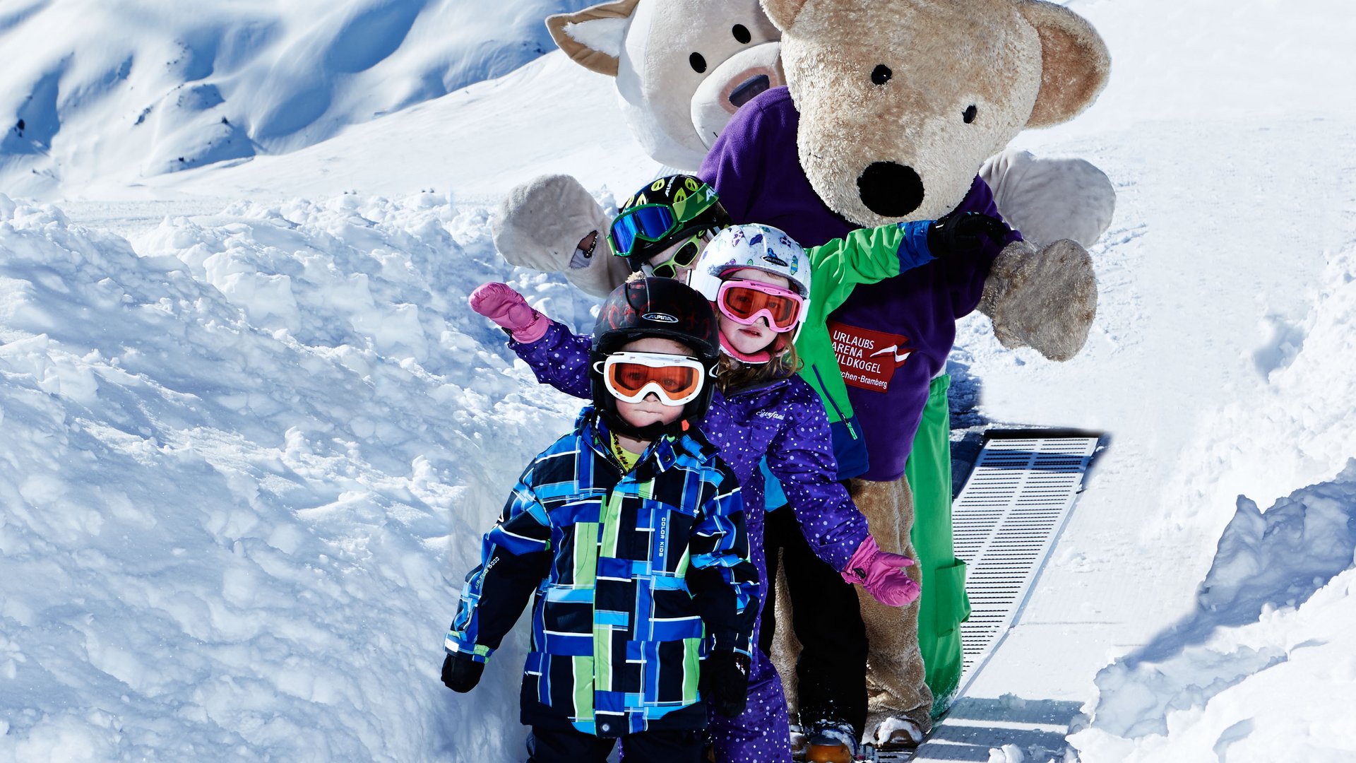 Skiurlaub mit Kindern in Österreich Kinder und zwei Figuren in Kostümen stehen auf einem Förderband in einer schneebedeckten Berglandschaft