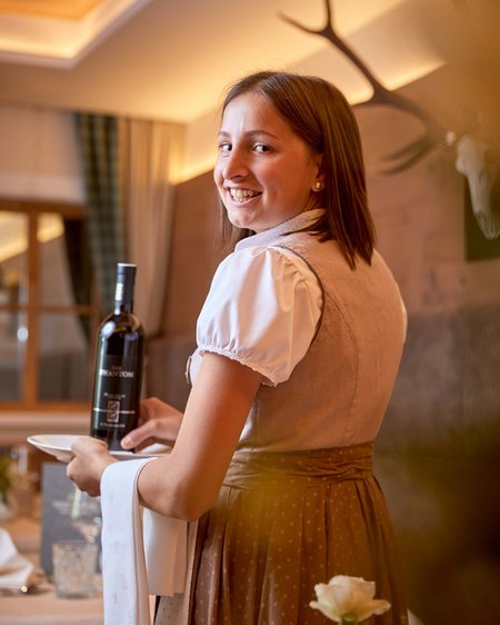 Langlaufurlaub Österreich: Smaragdhotel Tauernblick Lächelnde Kellnerin in traditioneller Kleidung hält Weinflasche und Teller im Restaurant