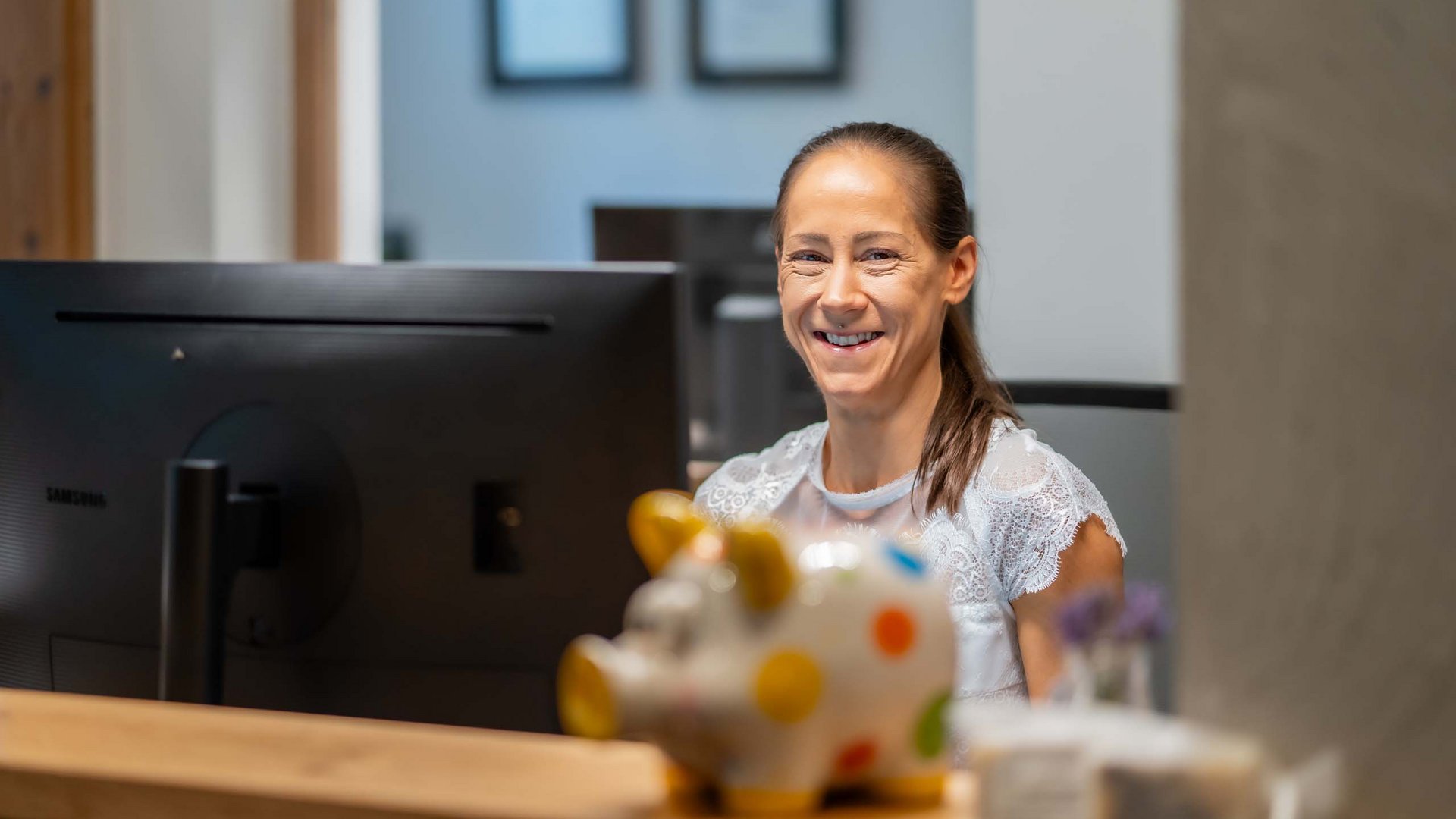 Wildkogel: Hotel mit Blick für das Besondere Lächelnde Frau am Empfangstresen mit Computer und bunter Sparschweinfigur