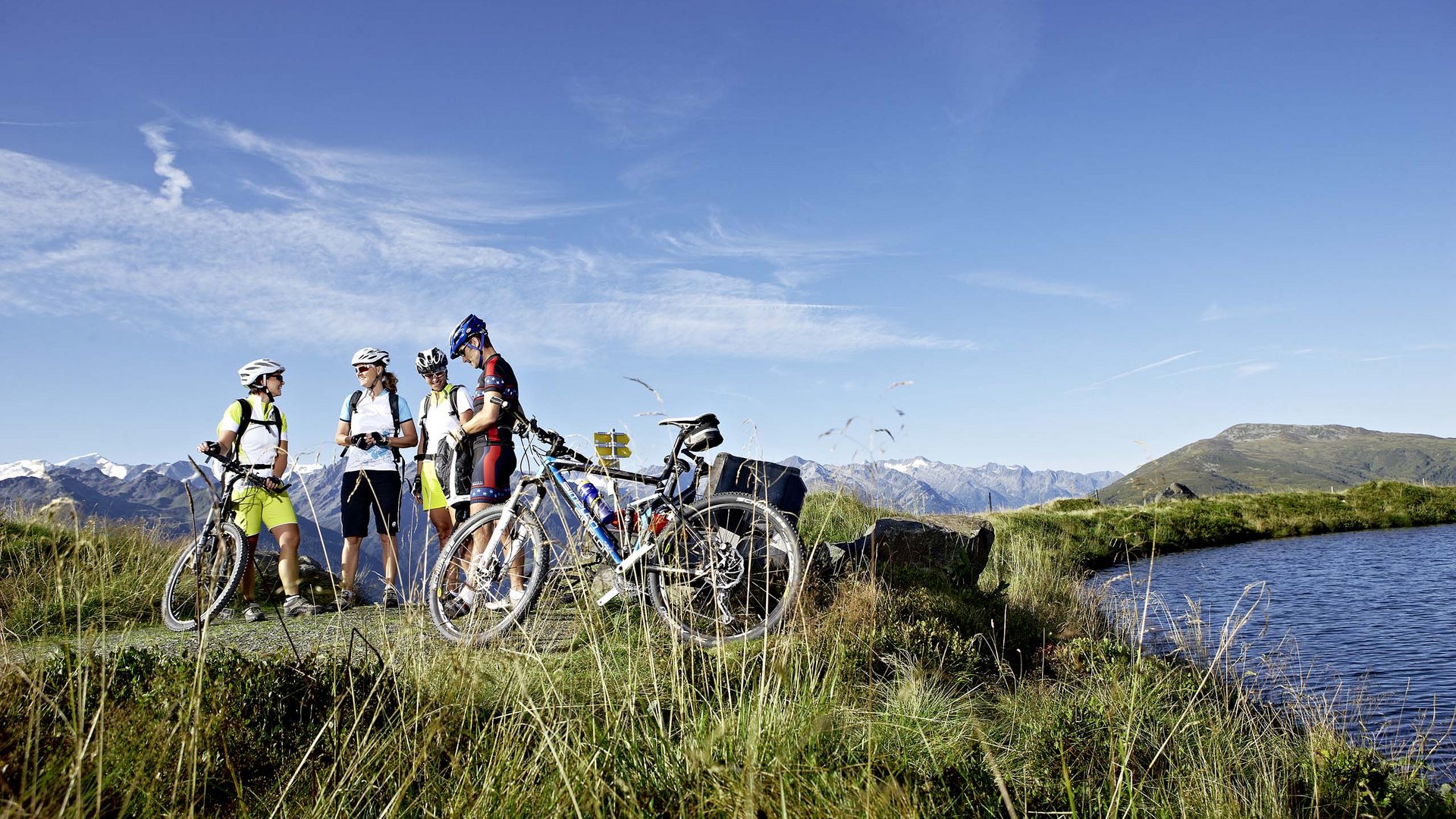 Hohe Tauern National Park: holiday for bikers Four cyclists with mountain bikes by a mountain lake under blue sky