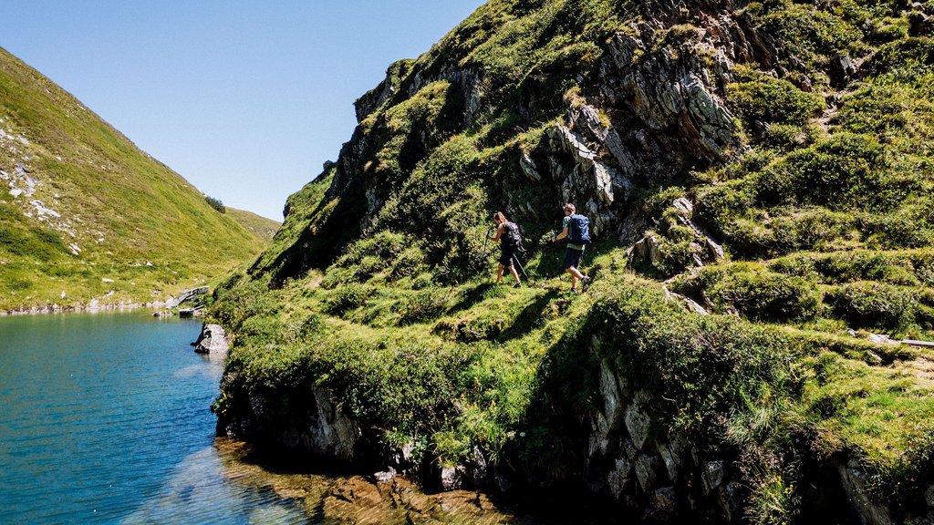 4 Sterne Hotel in Salzburgerland: Smaragdhotel Tauernblick Zwei Wanderer auf einem grasbewachsenen Pfad neben einem klaren Bergsee an einem sonnigen Tag