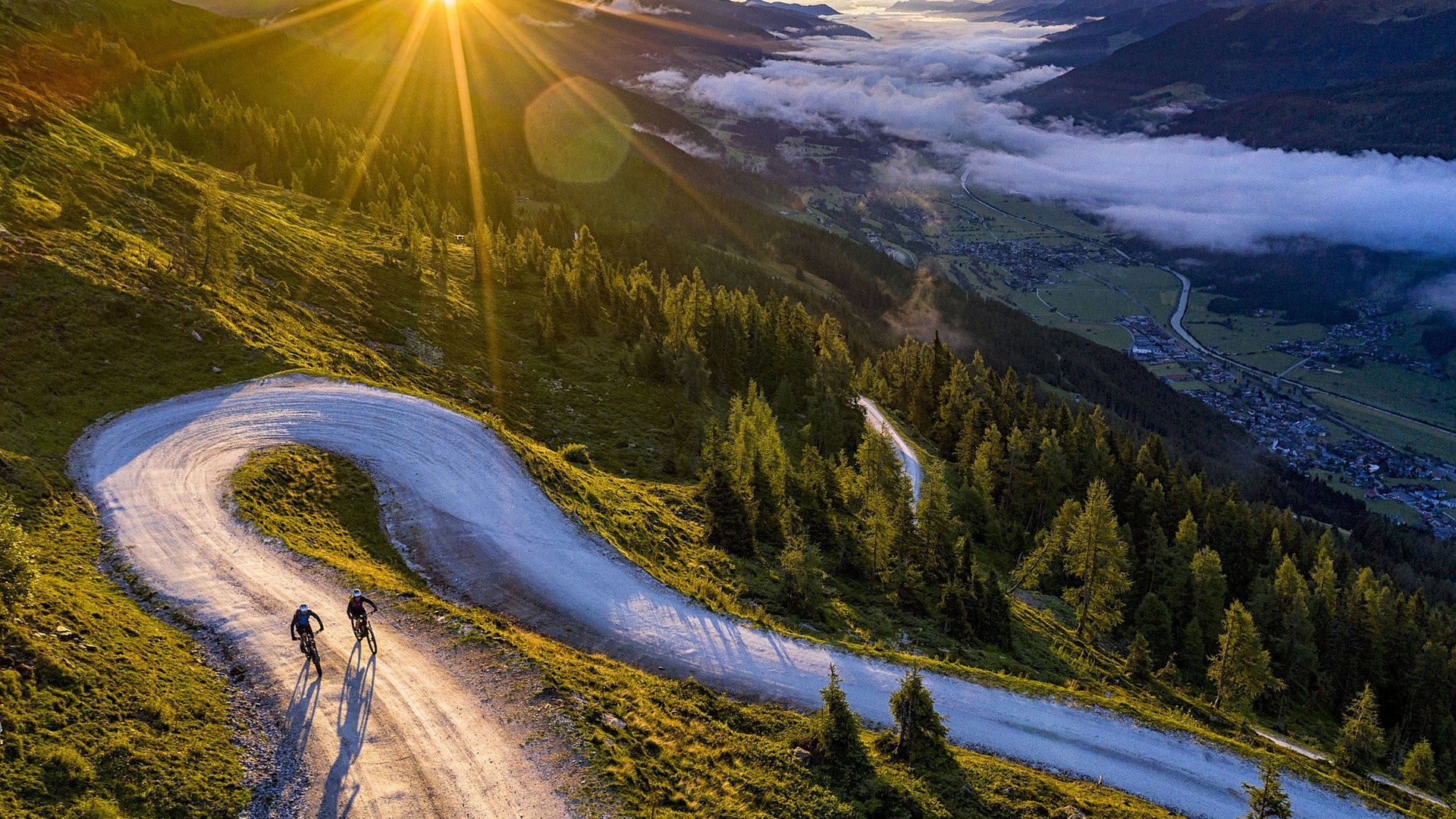 Familienhotel: Österreich im Sommer erleben Zwei Radfahrer auf einem kurvigen Bergweg bei Sonnenaufgang mit Blick auf Tal und Wolken