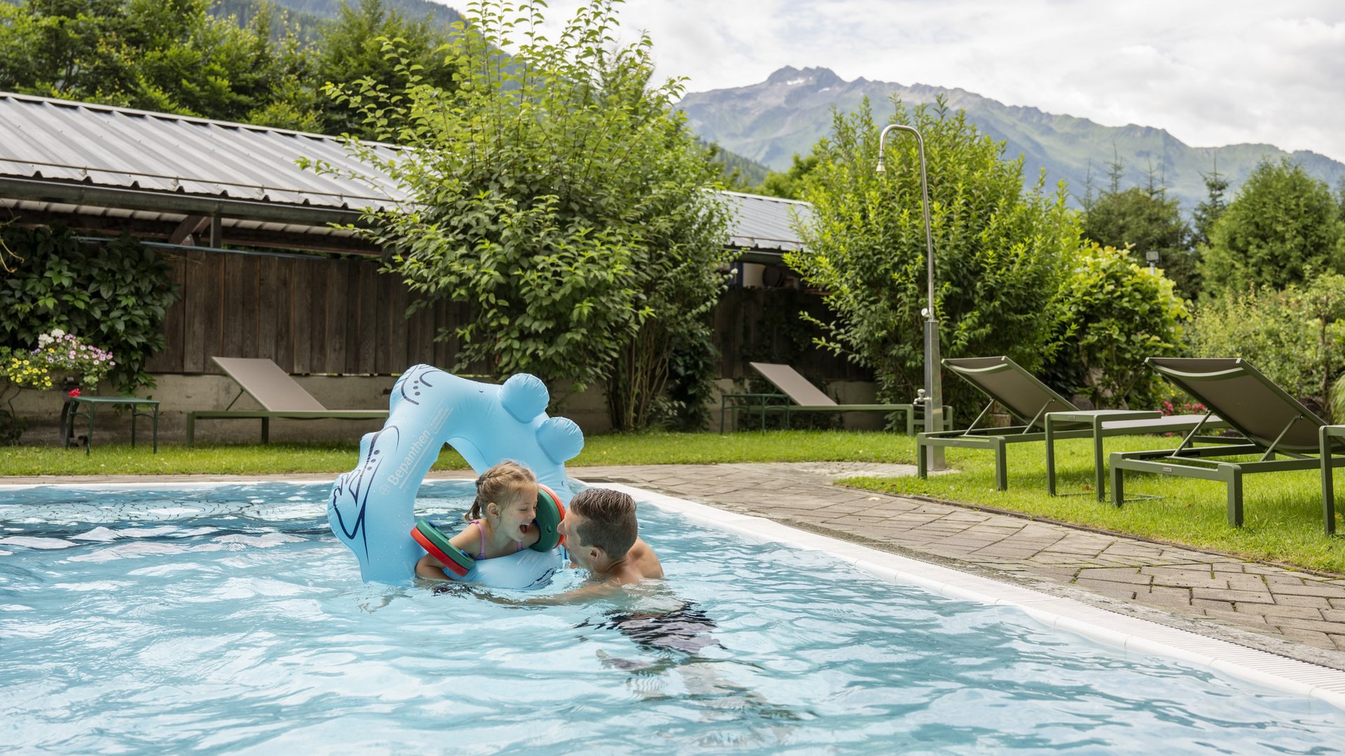 Familienhotel in Österreich mit Pool Mann spielt mit Kind im Pool mit Bergblick und Liegestühlen im Garten
