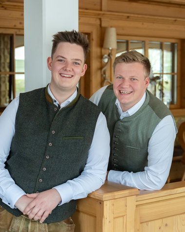 Wildkogel: Hotel mit Blick für das Besondere Zwei junge Männer in traditionellen Trachtenposen in einer hölzernen Innenraumumgebung