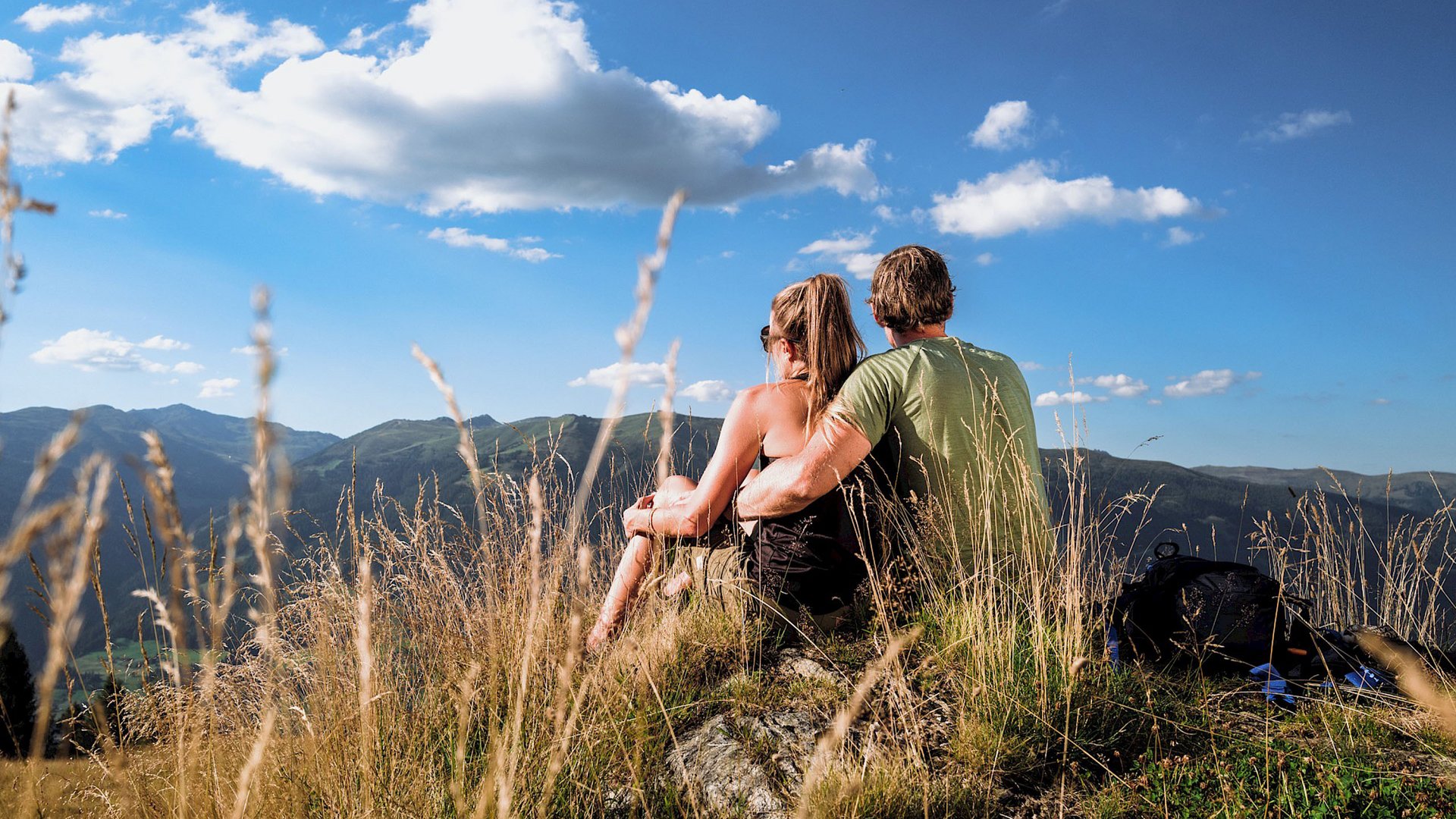 Familienhotel: Österreich im Sommer erleben Paar sitzt auf Berg mit Rucksack und schaut auf Landschaft unter blauem Himmel