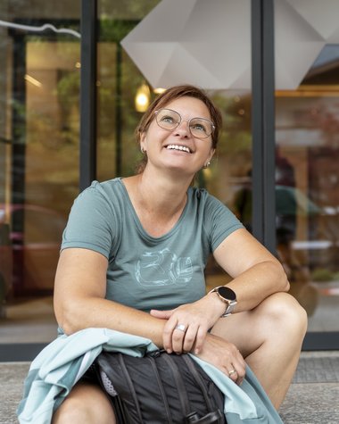 Wildkogel: Hotel mit Blick für das Besondere Lächelnde Frau mit Brille sitzt draußen mit Rucksack und Wasserflasche