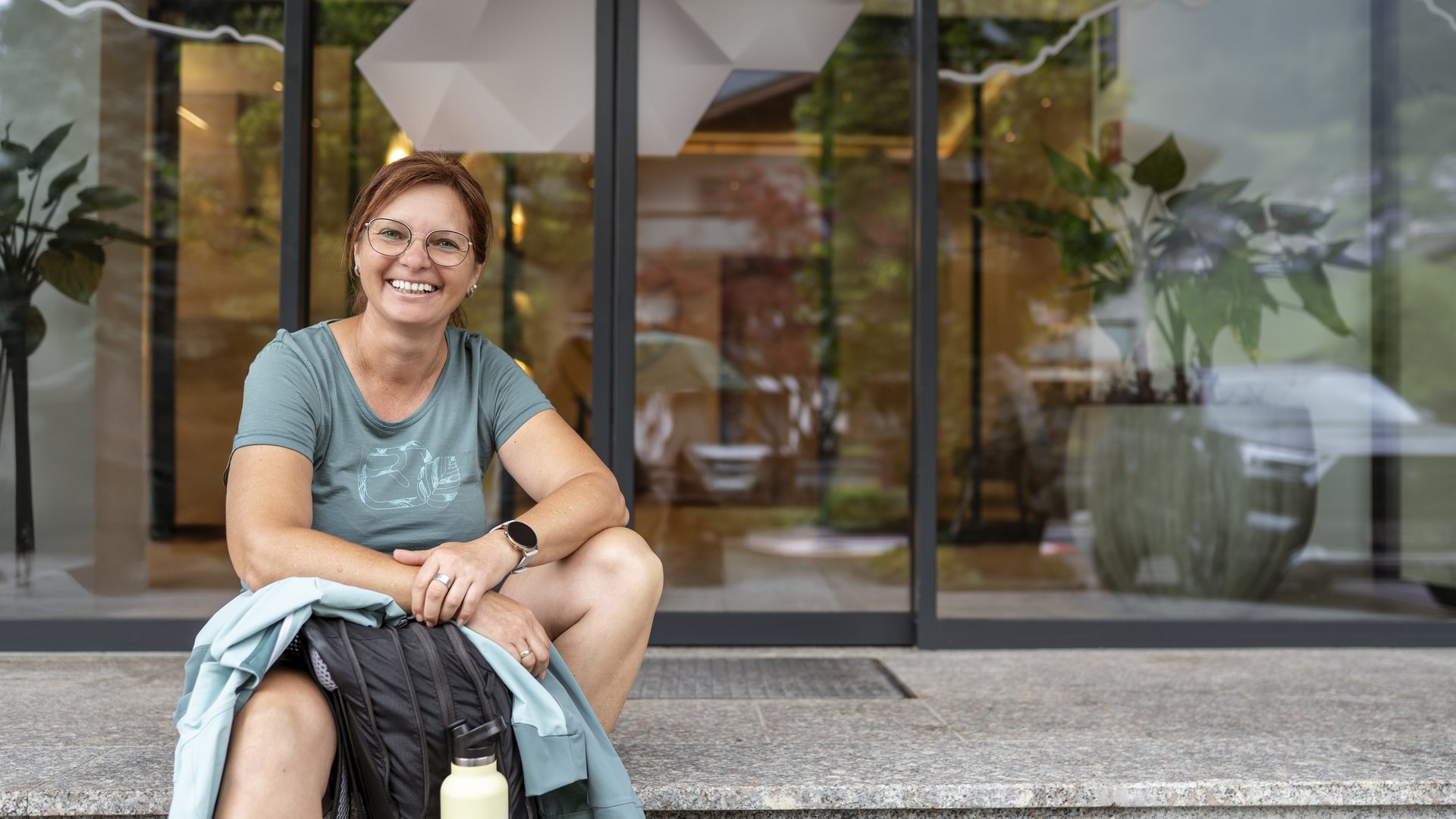 Wildkogel: Hotel mit Blick für das Besondere Fröhliche Frau mit Brille sitzt vor einem Gebäude mit Rucksack und Trinkflasche